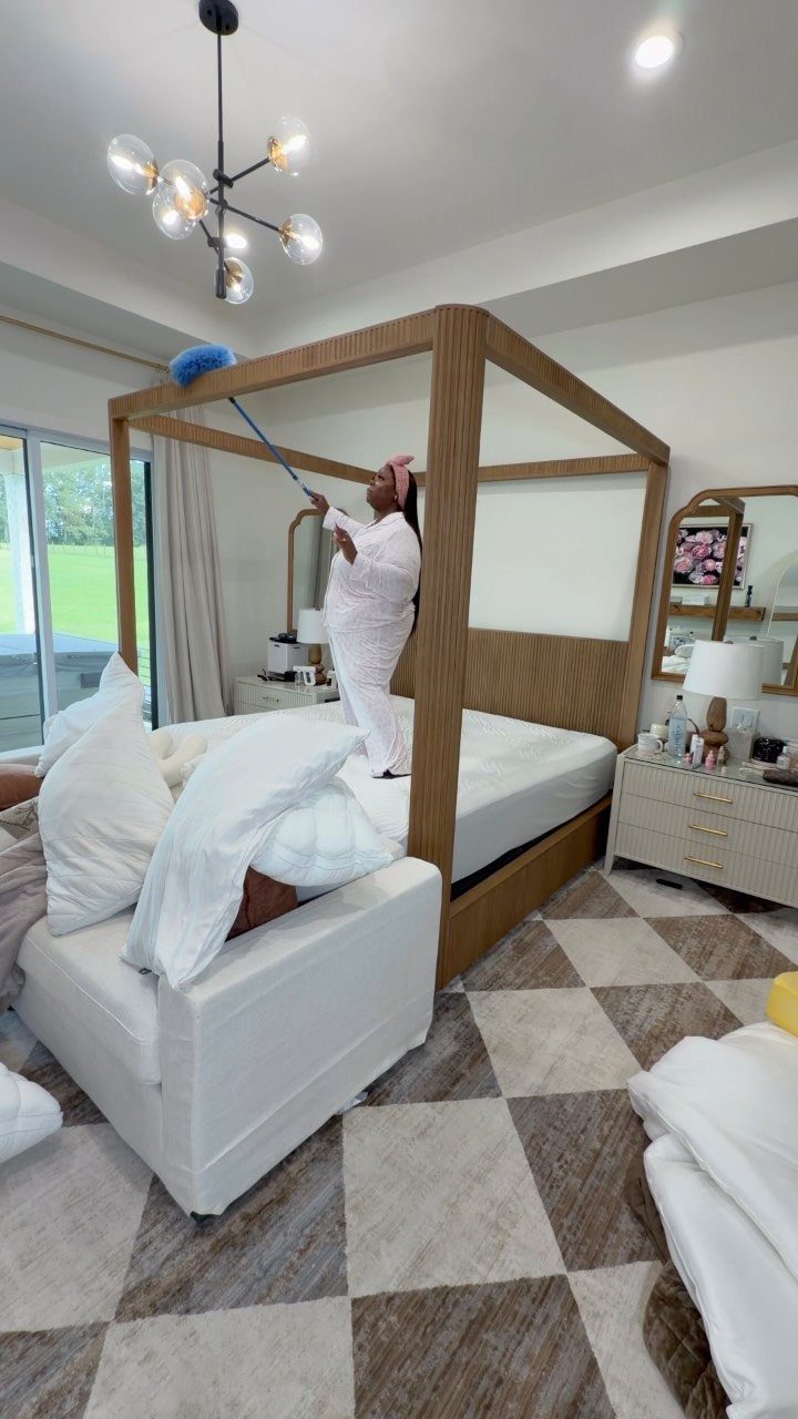 Apartment Deep Clean: 14 Areas to Scrub, Dust, and Vacuum 2 Person dusting the top of a wooden four-poster bed in a bright bedroom.