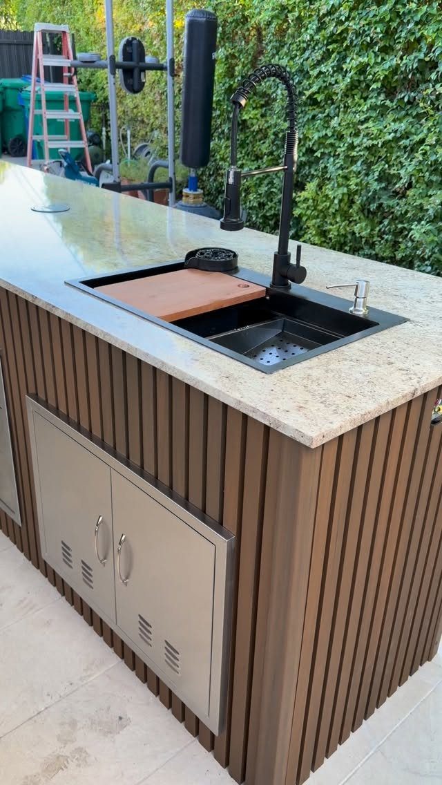 Outdoor kitchen island with granite countertop, black sink, and brown slatted base.