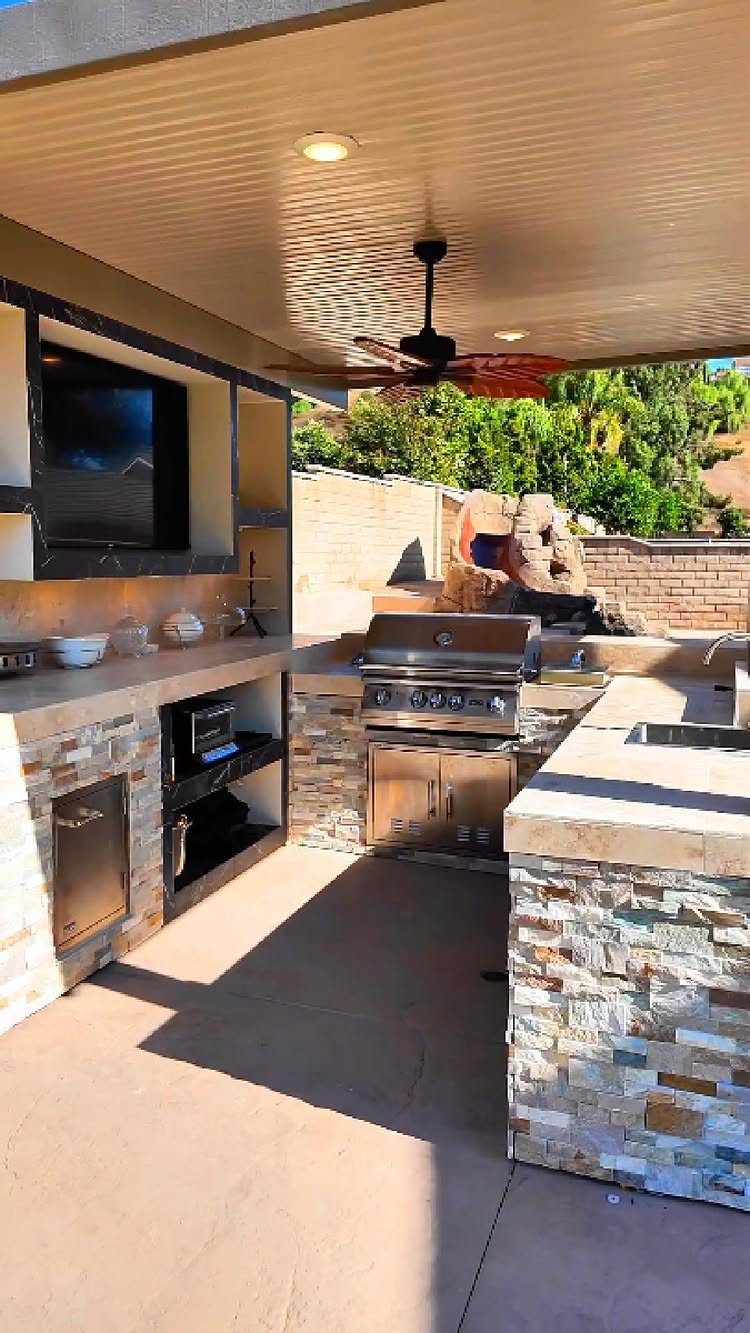 L-shaped outdoor kitchen with stone cladding, built-in grill, and mounted television under patio.