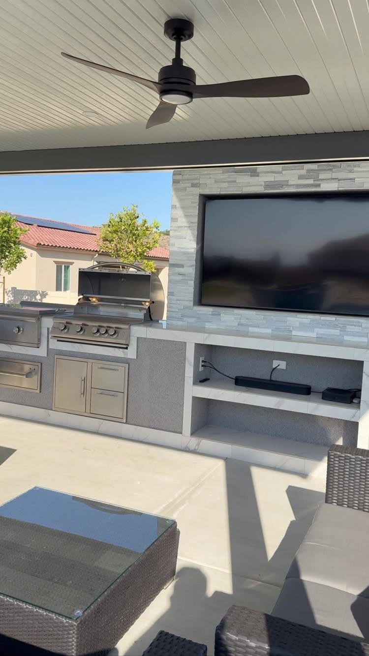 Outdoor kitchen with stainless steel grill and television mounted on a stone wall.