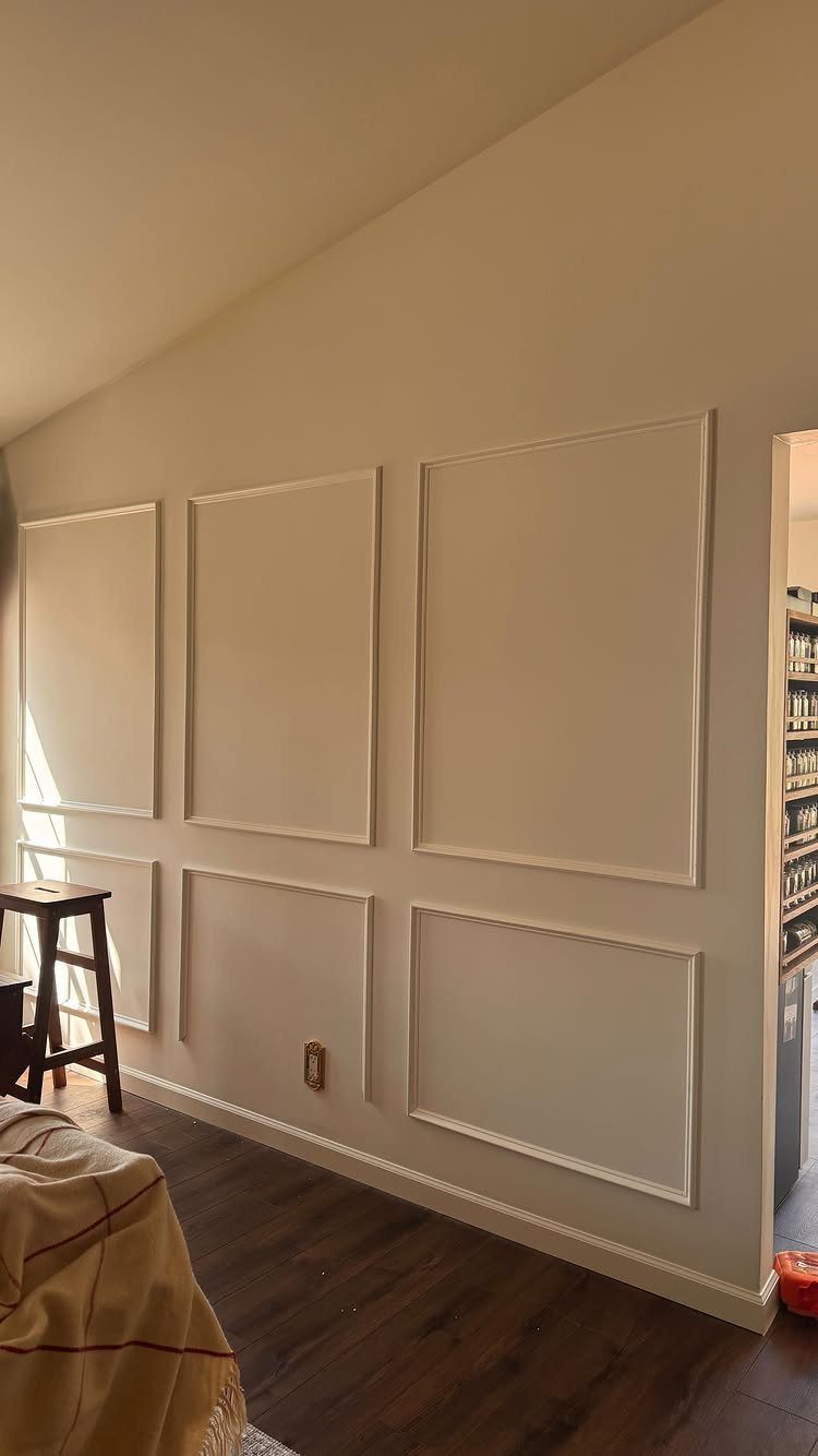 Cream colored wall decorated with large rectangular box moulding panels and dark wood flooring.
