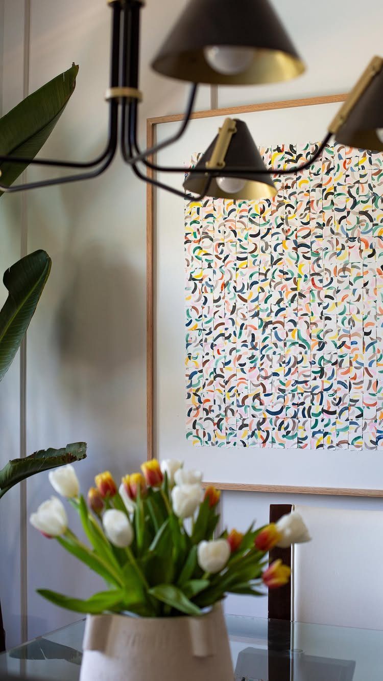 Framed abstract painting with colorful curved strokes hanging on a wall behind a lamp and tulips.