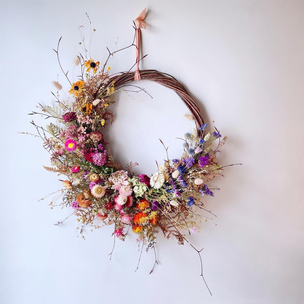 A colorful dried flower wreath made of twigs hanging on a white wall.