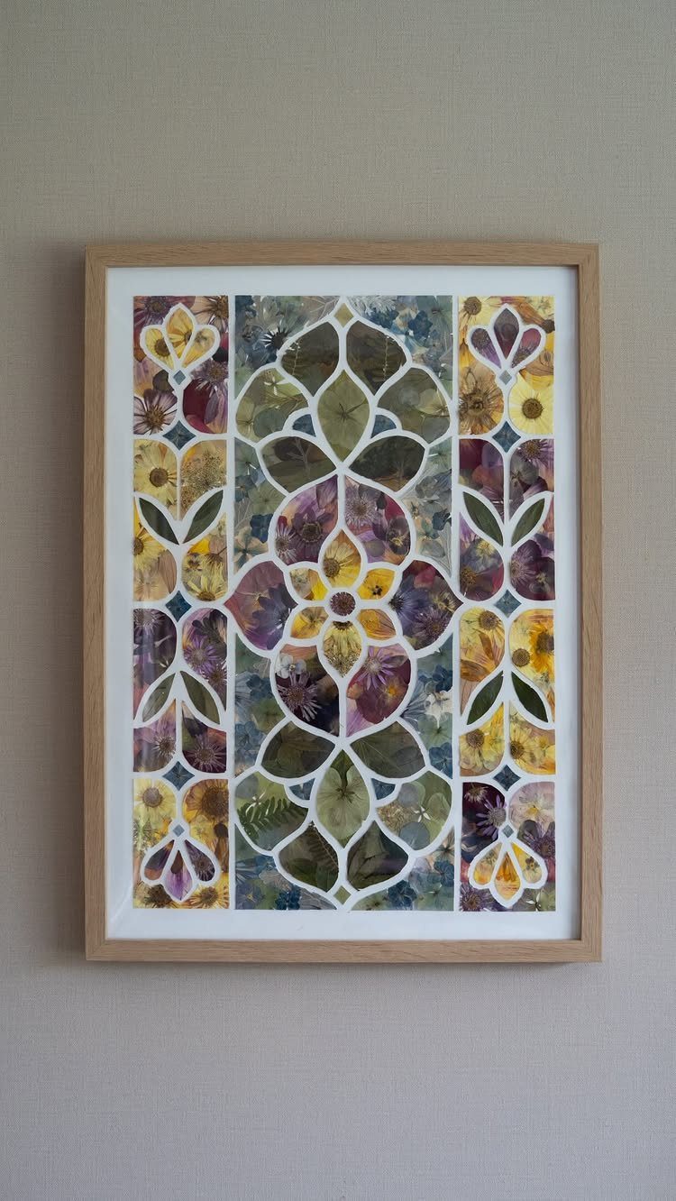 Pressed flower art in a wooden frame featuring a symmetrical botanical pattern on a neutral wall.