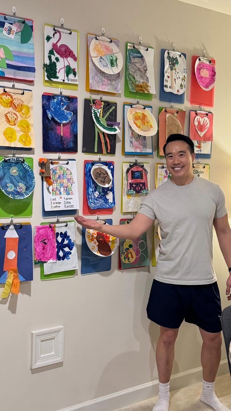 A smiling man points to colorful children's drawings clipped to a wall.