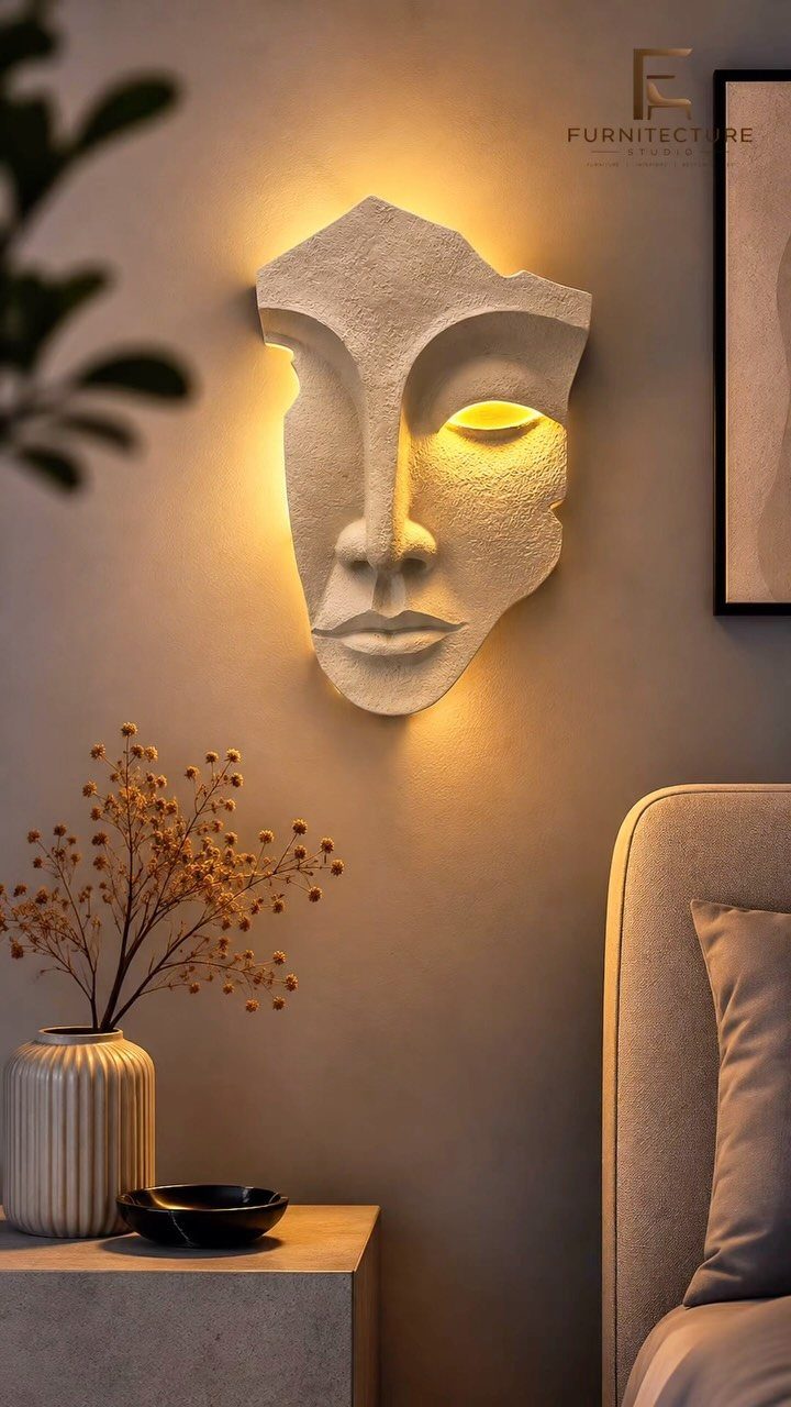A white textured face sculpture wall lamp glowing with warm light against a beige wall.