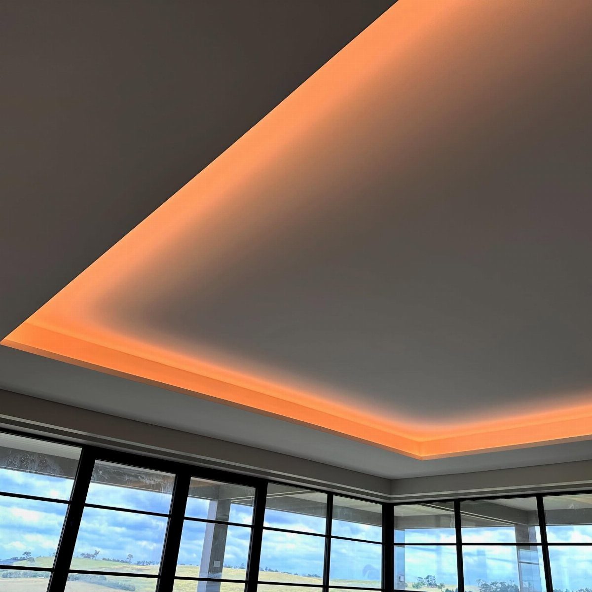 White recessed tray ceiling with warm orange cove lighting and large black-framed windows.