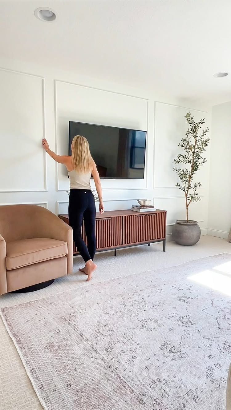 Woman touching white rectangular picture frame molding on a living room wall with a television.