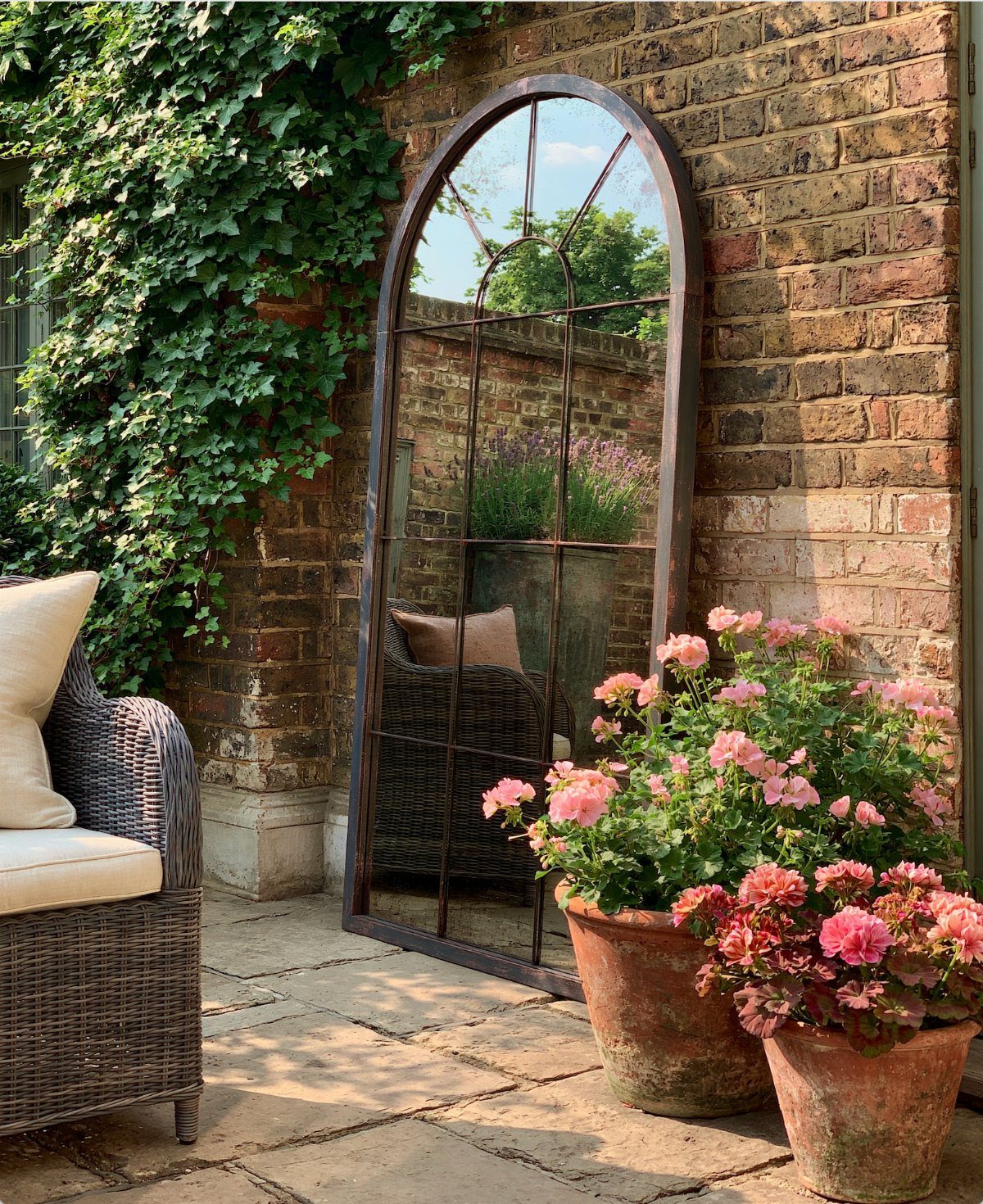 20 Garden Ideas for Small Spaces, From Vertical to Fire Pits 9 Arched mirror leaning against a brick wall reflecting a sunny garden with pink flowers.