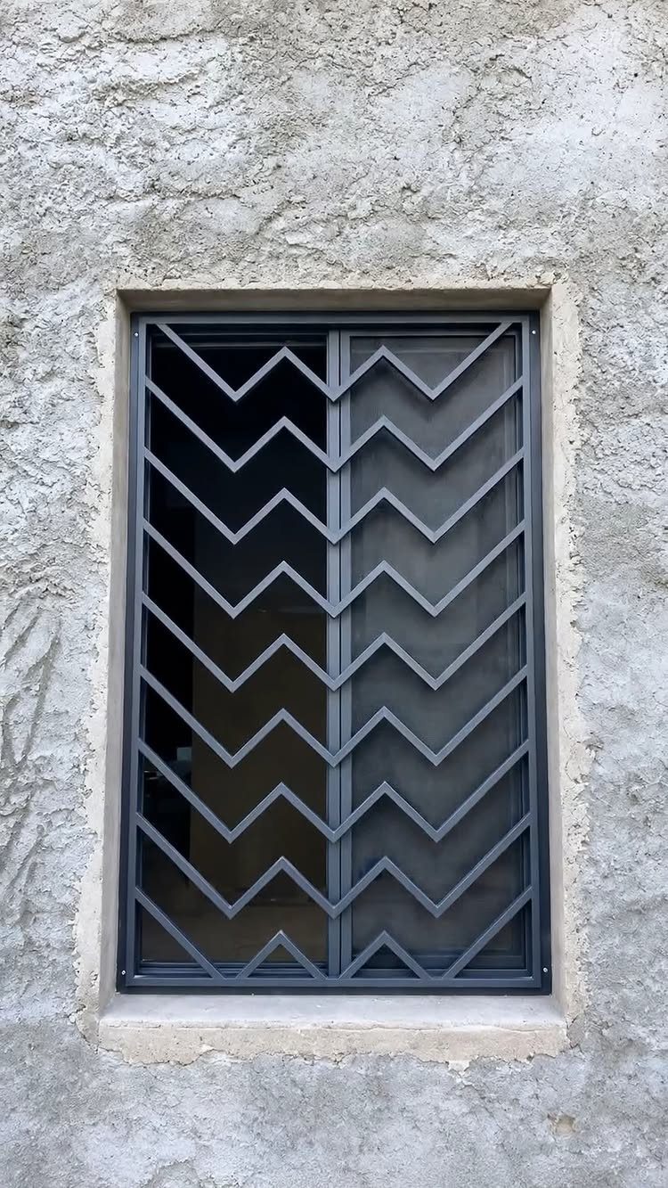 Window with a dark grey metal security grill in a zigzag pattern on concrete.