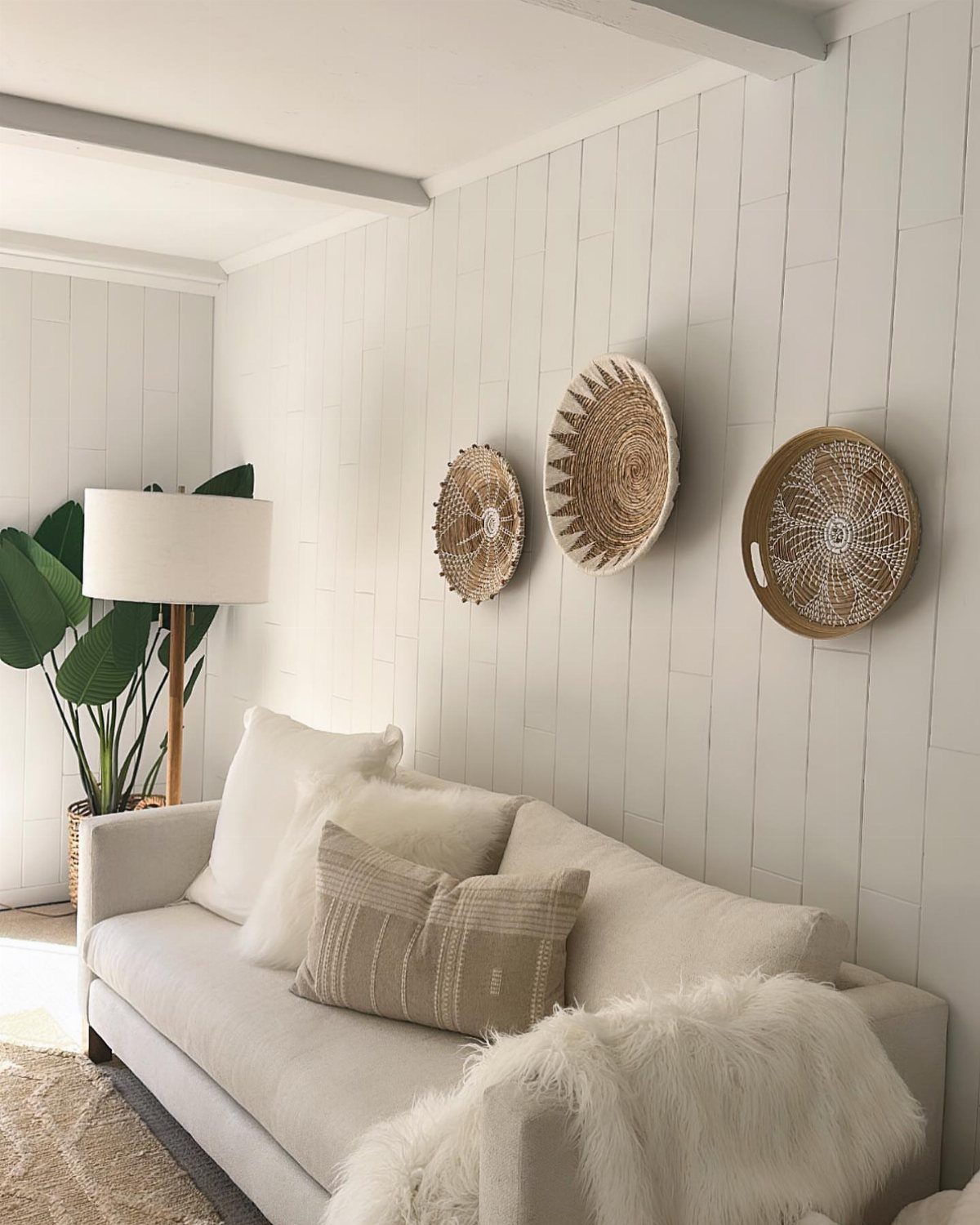 White vertical wood paneled wall with three woven baskets hanging above a neutral sofa.
