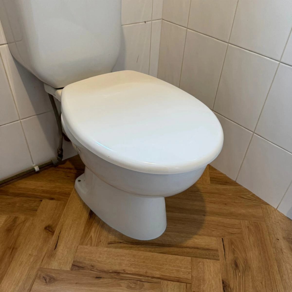 White toilet on wood-patterned herringbone flooring against white tiled bathroom walls.