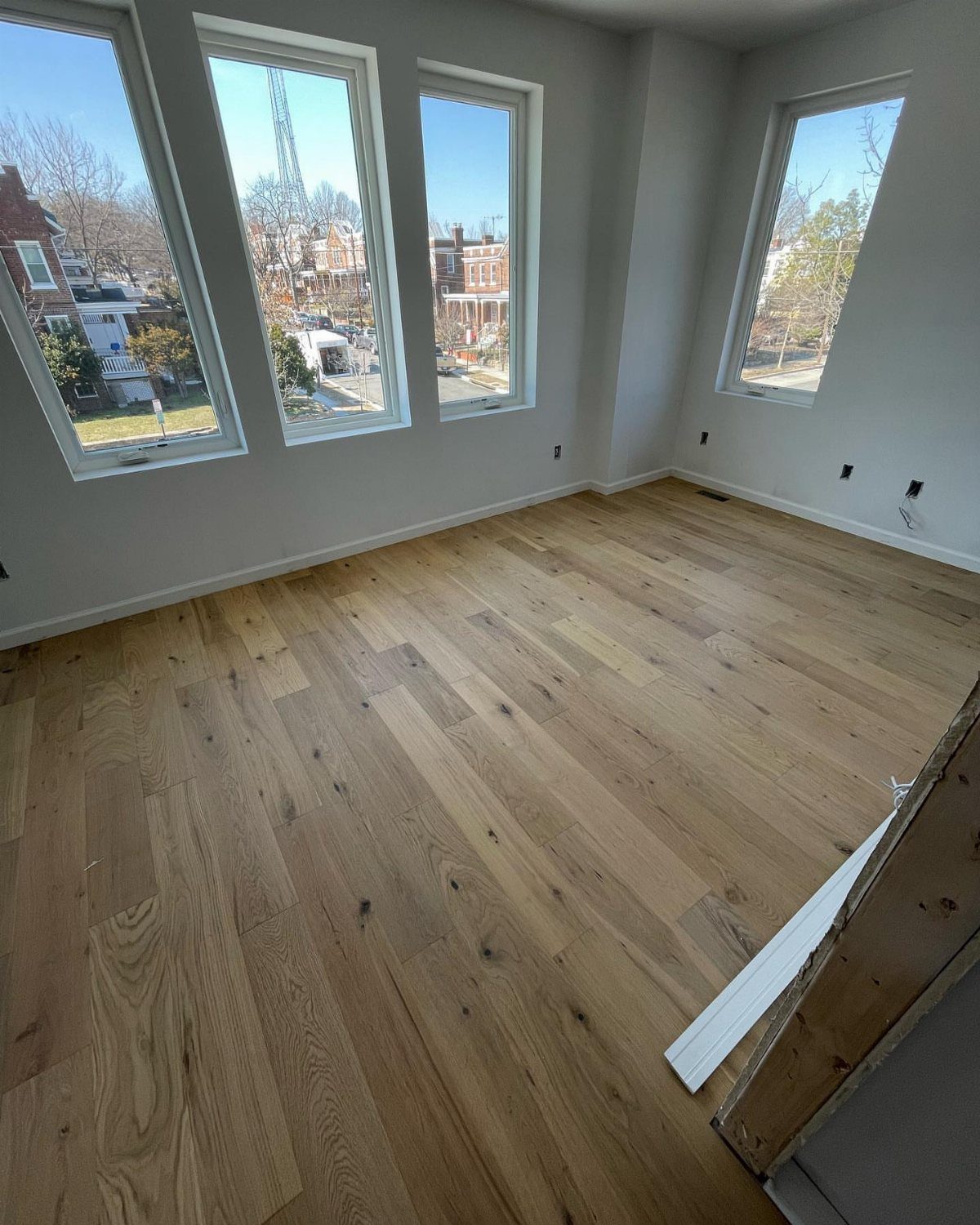 Medium Hardwood Floors: 5 Rooms That Get the Tone Right 2 Medium-toned hardwood flooring installed in an empty room with large windows overlooking a neighborhood.