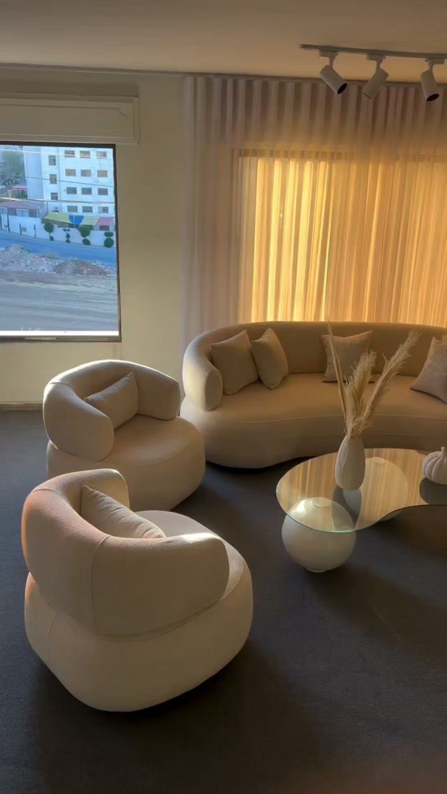 Curved beige sofa and matching armchairs arranged around a glass coffee table in a minimalist living room.