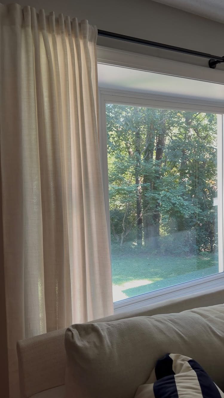 Cream sheer curtains hanging by a large window overlooking a lush green garden.