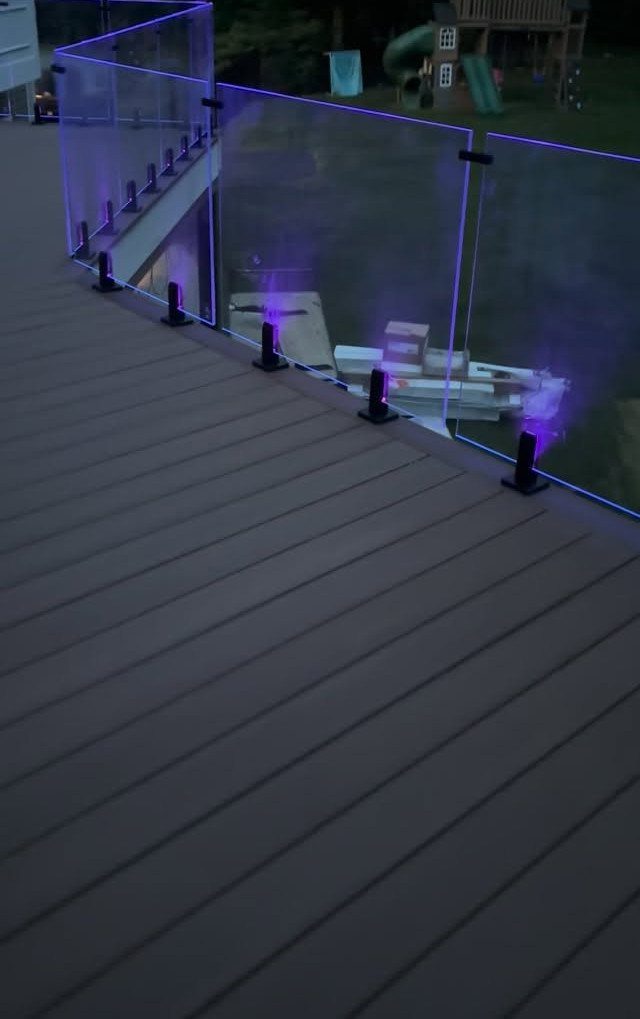 25 Patio Lights Ideas, From String Lights to Step Lighting 22 Wooden deck with glass railing illuminated by small purple LED lights at the base.