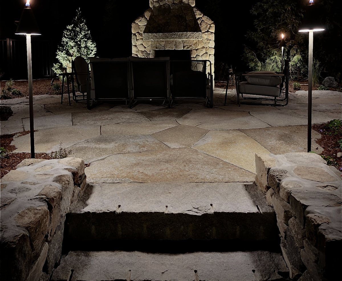 25 Patio Lights Ideas, From String Lights to Step Lighting 8 Stone outdoor fireplace and torch lights on a flagstone patio at night.