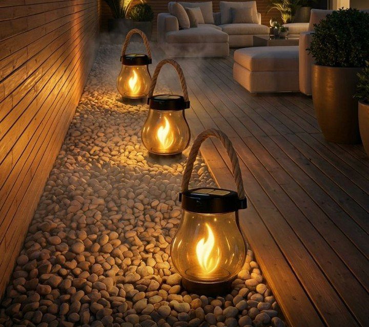 25 Patio Lights Ideas, From String Lights to Step Lighting 3 Three glowing glass lanterns with rope handles sitting on a pebble path by a deck.