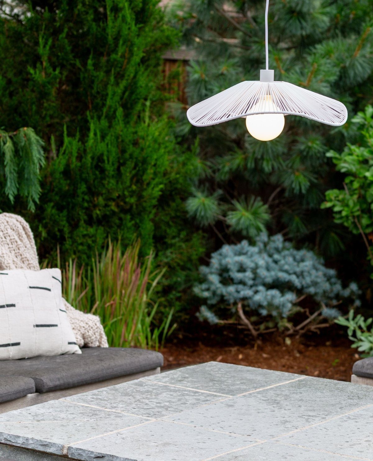 25 Patio Lights Ideas, From String Lights to Step Lighting 14 White pendant light hanging over a stone patio with green garden foliage.