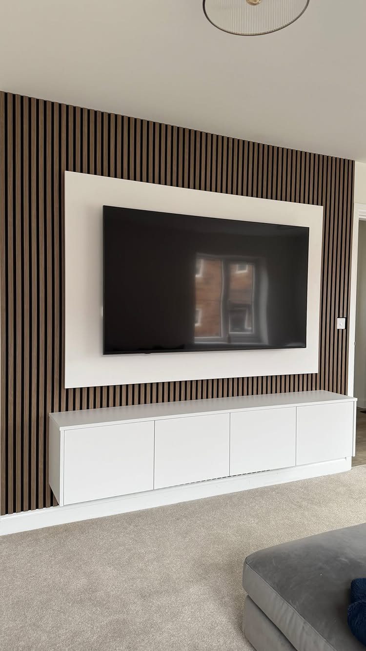 A television mounted on a white panel against a dark wood slat wall with a white console.