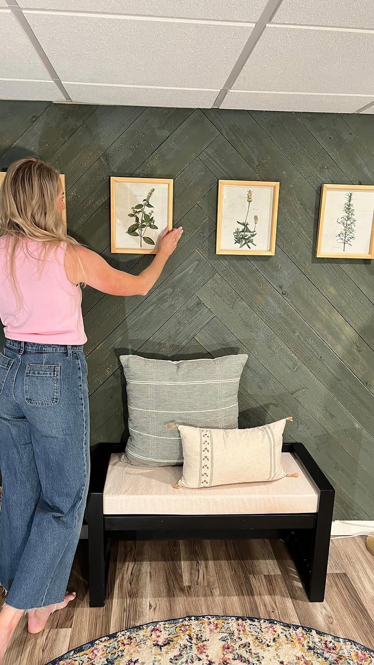 Woman hanging a framed botanical print on a dark green diagonal planked accent wall.