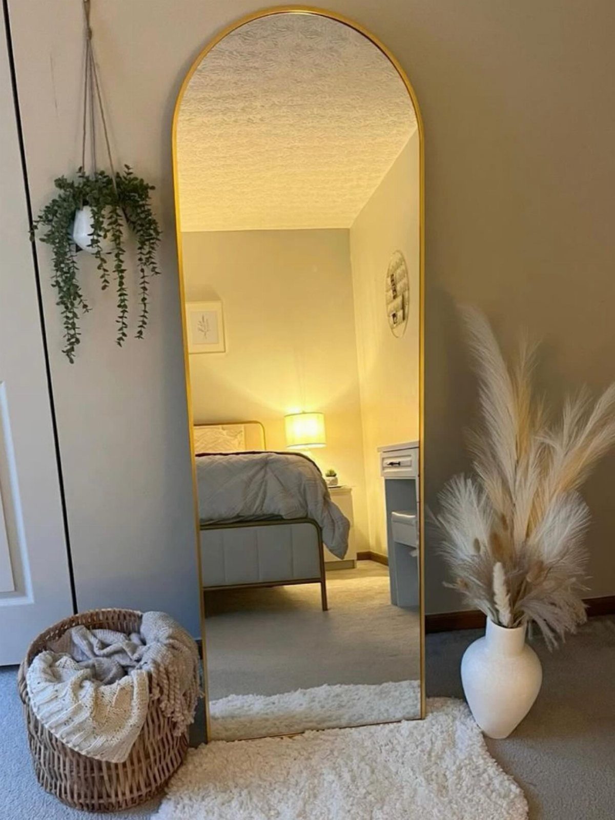 Arched gold-framed mirror leaning against a wall reflecting a cozy bedroom interior