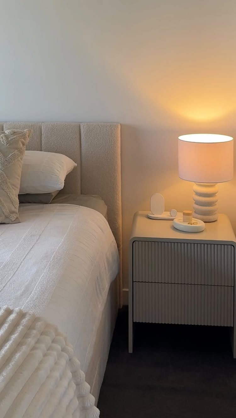 White bedside table with a glowing ribbed lamp and decorative tray next to a bed.