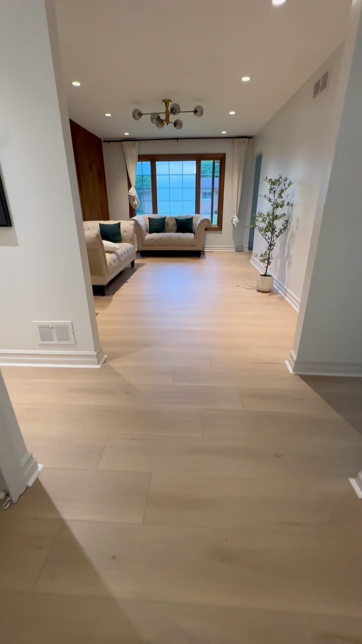 Light wood plank flooring leading into a bright living room with a beige sofa and large window.